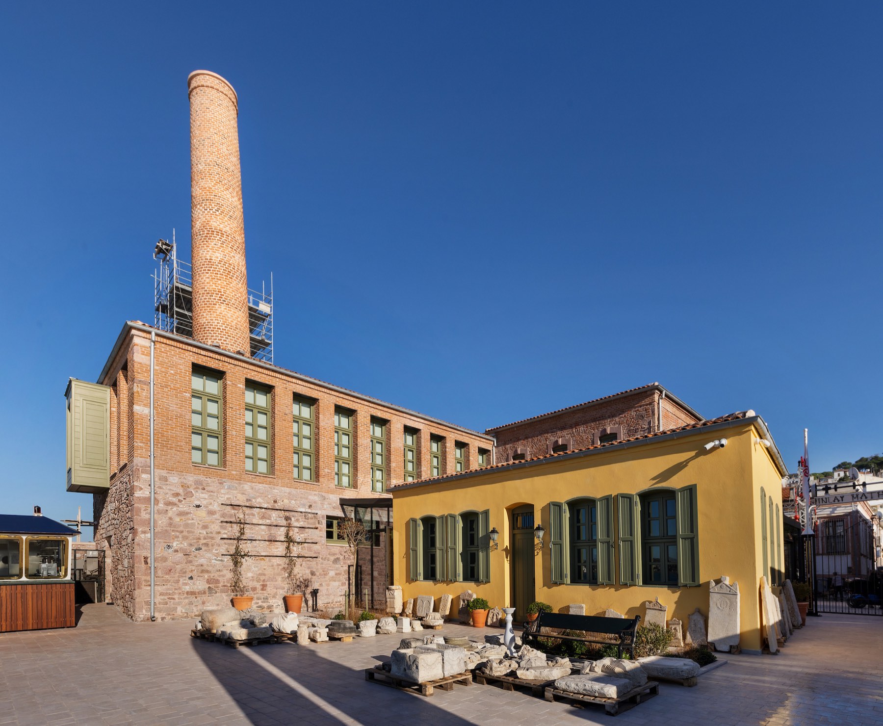 Avludan bakış: taş eserler sergilenirken arkada fabrika cephesi ve baca